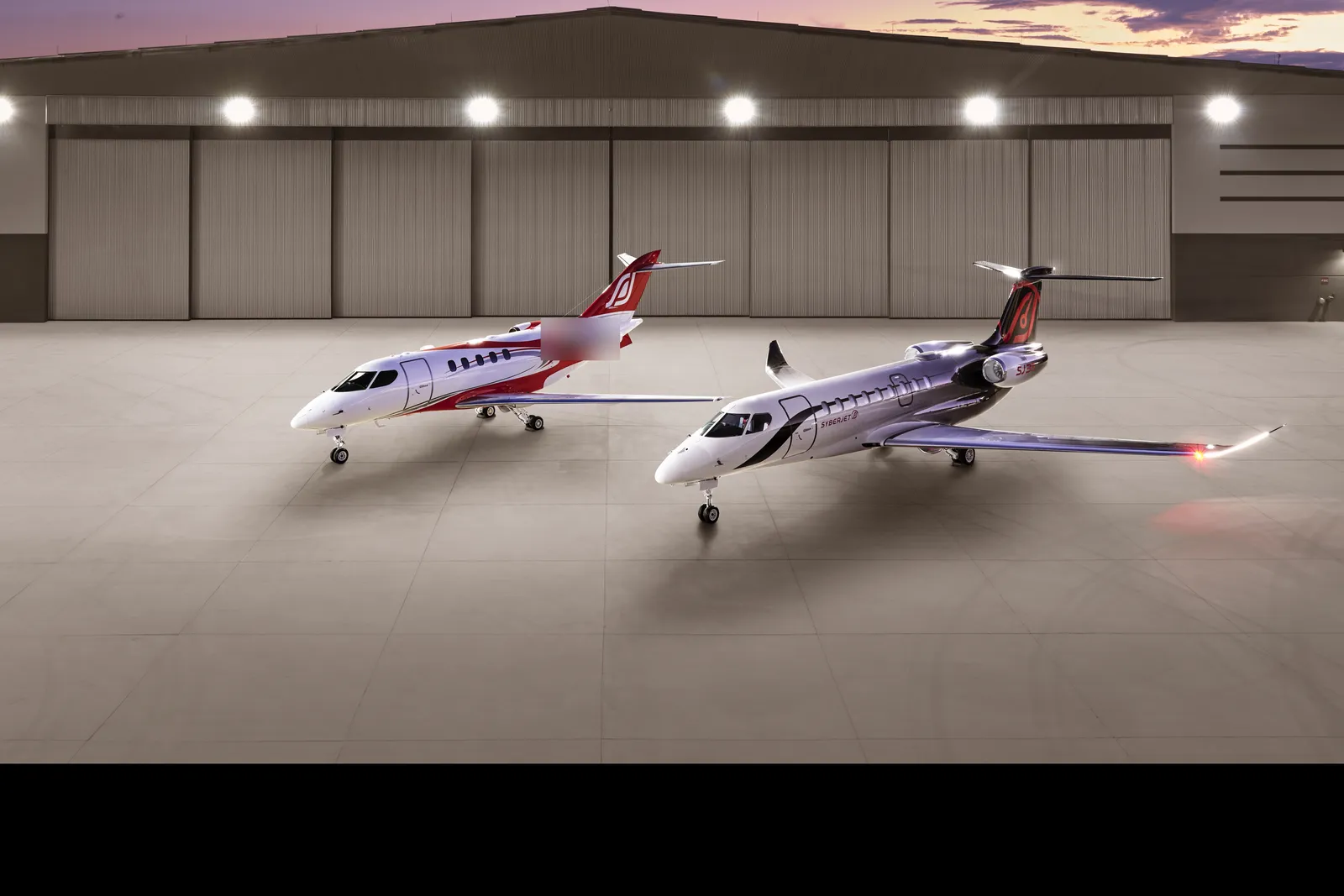 Red and white SyberJet SJ30 parked next to the silver SJ36 in front of a hangar at dusk
